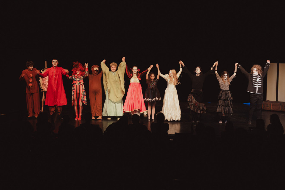 Eine Gruppe von Schauspielerinnen und Schauspielern in bunten Kostümen steht auf einer dunklen Theaterbühne und hält sich an den Händen. Vor ihnen sind die Silhouetten des Publikums zu sehen, während die Darstellenden offenbar Applaus entgegennehmen.