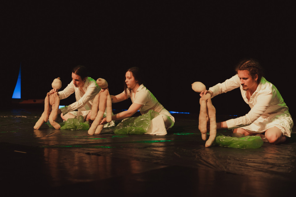 Auf einer dunklen Bühne sitzen drei weiß gekleidete Personen und bewegen weiche, menschenähnliche Puppen. Grüne Folie auf dem Boden und farbiges Licht verleihen der Szene eine experimentelle Theateratmosphäre.