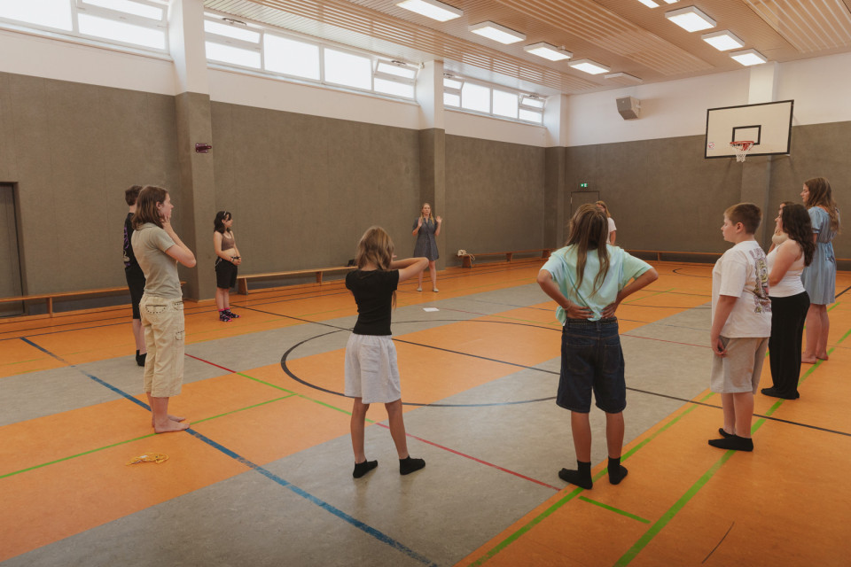 Eine Gruppe von Kindern steht in einer Sporthalle im Kreis und hört einer Frau in der Mitte zu, die offenbar Anweisungen gibt. Im Hintergrund sind ein Basketballkorb, hohe Fenster und die Linien des Hallenbodens zu sehen.