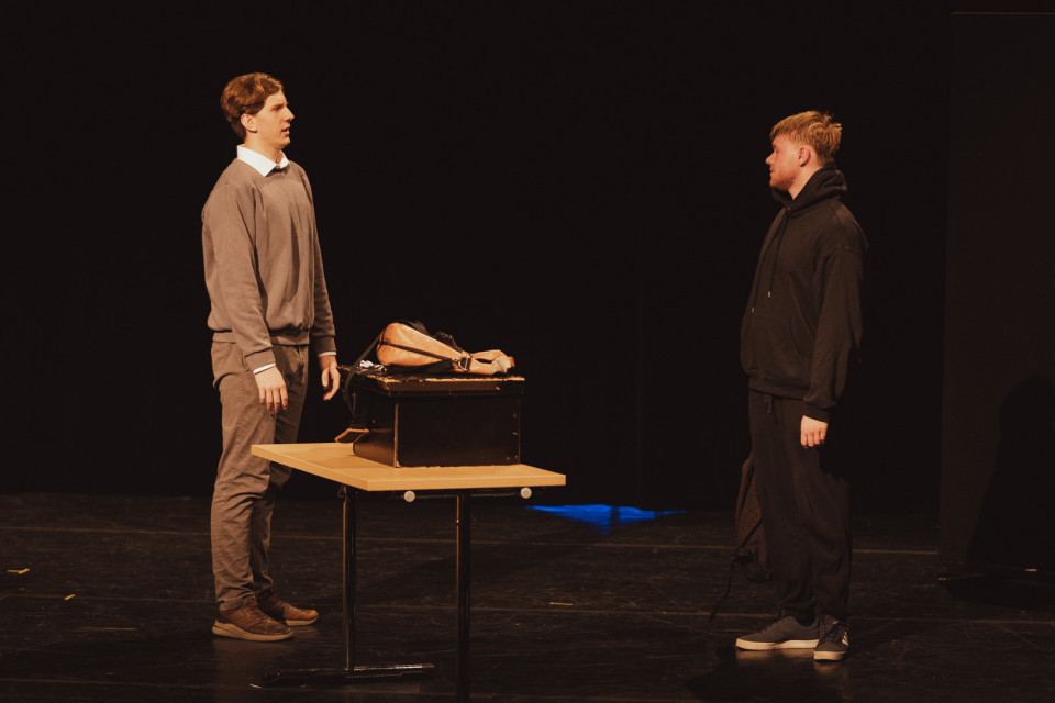 Auf einer dunklen Theaterbühne stehen sich zwei Männer gegenüber und scheinen miteinander zu sprechen. Zwischen ihnen befindet sich ein Tisch mit einem schwarzen Koffer, auf dem ein künstlicher Körperteil oder eine Puppe liegt.