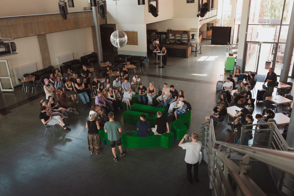 In einer großen, hellen Halle sitzen und stehen viele Menschen in einem Kreis um grüne Sofaelemente, offenbar bei einer Besprechung oder Veranstaltung. Im Hintergrund sind Tische, Stühle, eine Bar, große Fenster und eine Discokugel zu sehen.