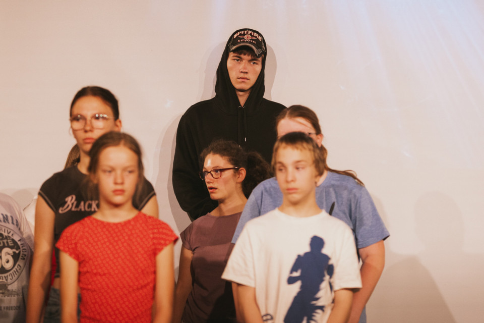Eine Gruppe von Kindern und Jugendlichen steht vor einer hellen Wand, wobei einige im Vordergrund unscharf zu sehen sind. Im Mittelpunkt steht ein Jugendlicher in schwarzem Hoodie und Baseballkappe, der ernst in die Kamera blickt.
