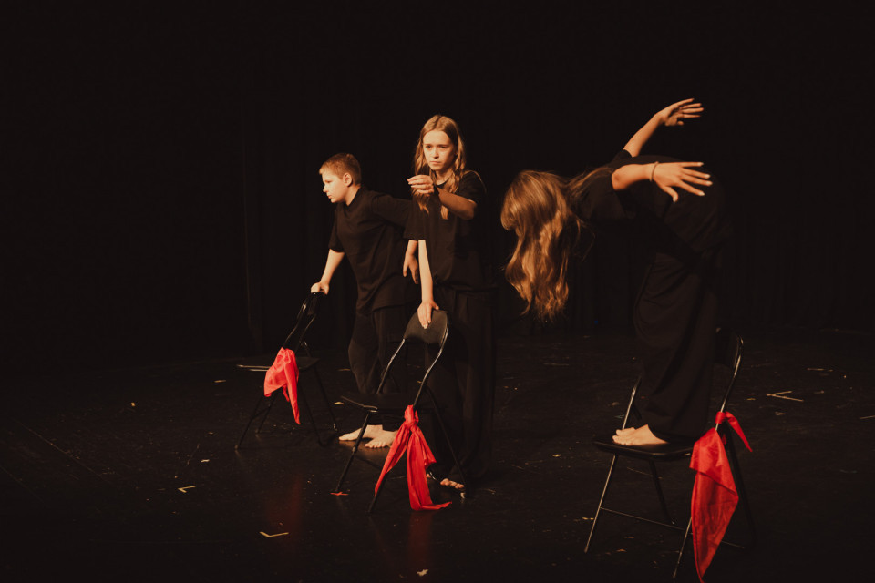 Drei Kinder in schwarzer Kleidung führen auf einer dunklen Bühne eine ausdrucksstarke Tanz- oder Theaterperformance mit Stühlen auf. An den Stühlen hängen rote Tücher, während die warm beleuchteten Figuren dynamische Posen einnehmen.