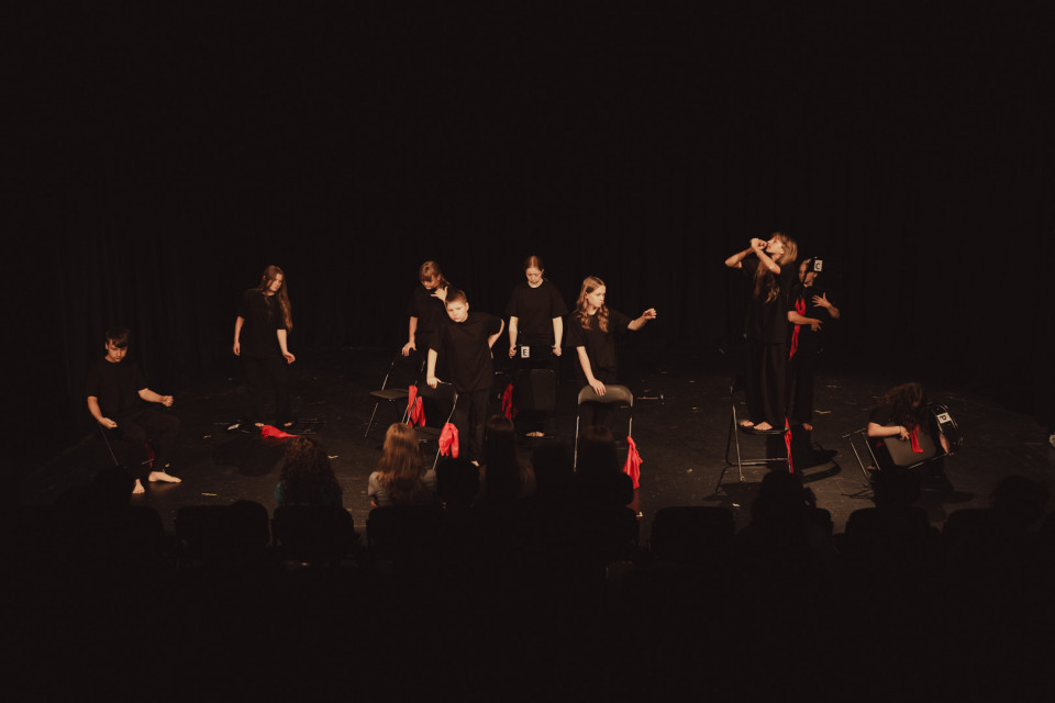 Auf einer dunklen Theaterbühne stehen und sitzen mehrere Kinder und Jugendliche in schwarzer Kleidung zwischen Stühlen mit roten Tüchern. Im Vordergrund sind die Silhouetten des Publikums zu sehen, während die Gruppe eine inszenierte Szene aufführt.