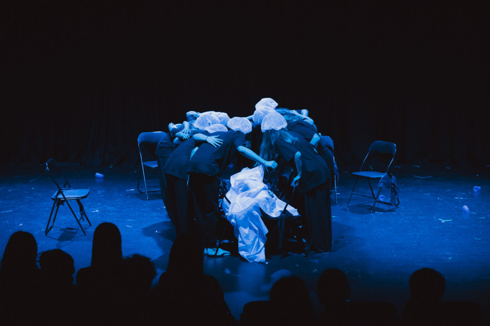 Auf einer dunkel beleuchteten Bühne kauert eine Gruppe von Darstellern in schwarzer Kleidung und weißen Hauben eng um mehrere Stühle. Blaues Licht, verstreute Requisiten und die Silhouetten des Publikums erzeugen eine dramatische Theateratmosphäre.