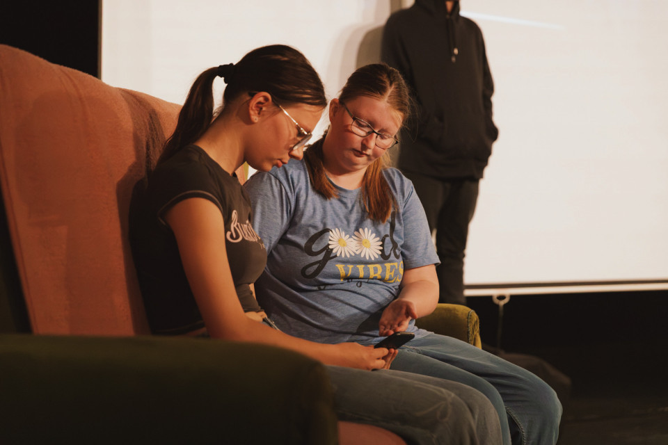 Zwei junge Frauen sitzen nebeneinander auf einem Sofa und schauen gemeinsam auf ein Smartphone. Im Hintergrund steht eine weitere Person in dunkler Kleidung vor einer hellen Wand oder Leinwand.