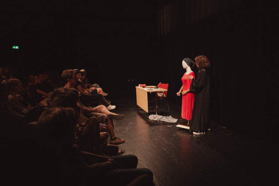 Auf einer dunklen Theaterbühne steht eine Person neben einer Schaufensterpuppe in einem roten Kleid, während links das Publikum zuschaut. Neben ihnen befinden sich ein kleiner Schreibtisch mit Stuhl und einige Requisiten im Scheinwerferlicht.