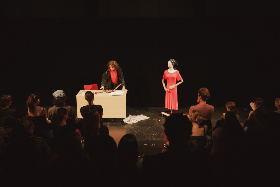Auf einer dunklen Theaterbühne steht eine Person hinter einem Tisch mit Papieren und Requisiten, während das Publikum zuschaut. Daneben steht eine Schaufensterpuppe in einem roten Kleid mit einem gemusterten Kopftuch im Scheinwerferlicht.