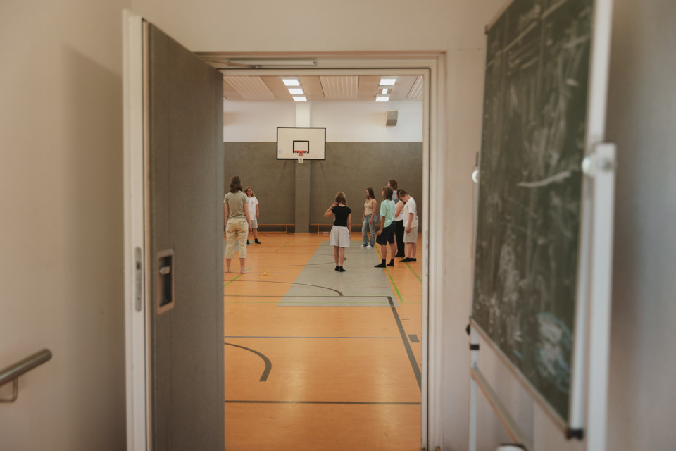 Durch eine offene Tür blickt man in eine Sporthalle, in der mehrere Jugendliche unter einem Basketballkorb stehen. Im Vordergrund sieht man einen Flur mit einer Tafel an der rechten Wand.