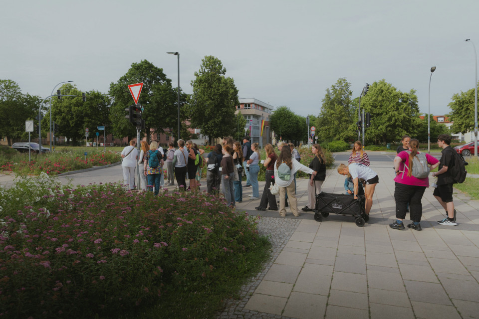 Eine Gruppe von Menschen steht an einer Kreuzung neben Blumenbeeten und wartet offenbar an der Ampel. Im Hintergrund sind Bäume, Verkehrsschilder, Straßenlaternen und einige Gebäude unter einem bewölkten Himmel zu sehen.