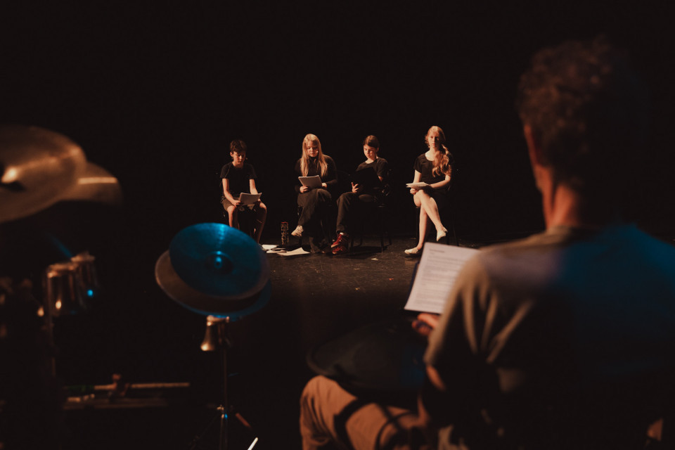Auf einer dunklen Bühne sitzen vier junge Personen in einer Reihe und lesen aus Papieren. Im Vordergrund ist unscharf eine weitere Person mit einem Blatt zu sehen, daneben stehen Schlagzeugbecken.