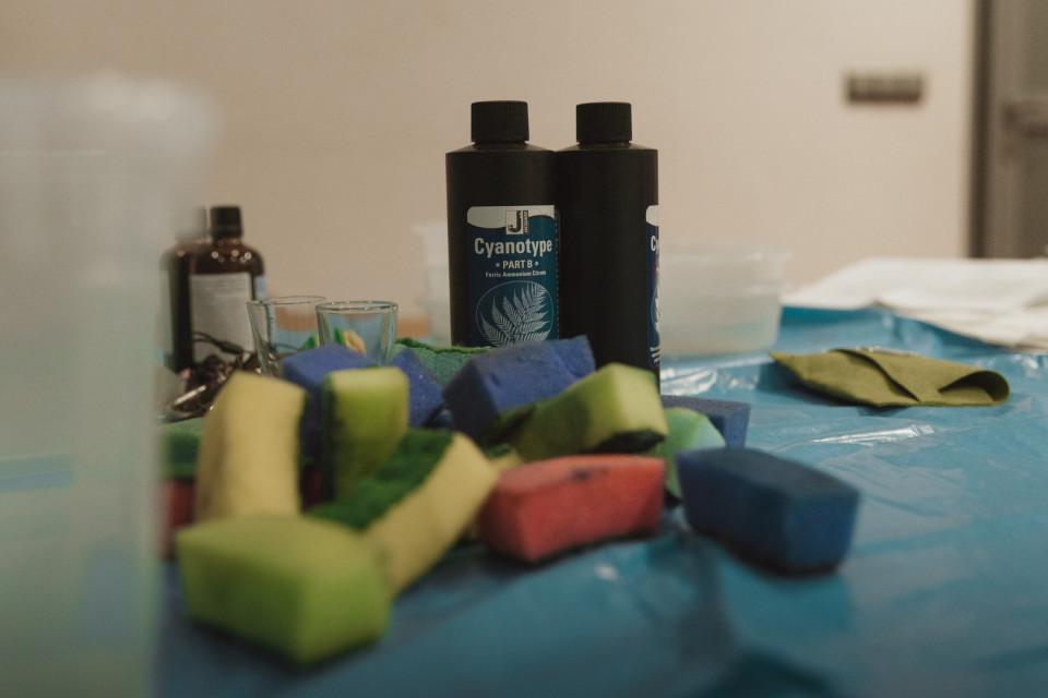 Auf einem mit blauer Folie bedeckten Tisch stehen zwei schwarze Flaschen mit der Aufschrift „Cyanotype“ zwischen kleinen Gläsern und weiteren Chemikalienbehältern. Im Vordergrund liegen unscharf mehrere bunte Schwämme und Stoffstücke, die wie Materialien für ein kreatives oder fotografisches Verfahren wirken.
