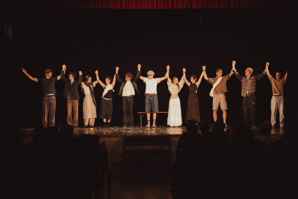 Eine Gruppe von Darstellerinnen und Darstellern steht auf einer Theaterbühne und hält sich nach einer Aufführung mit erhobenen Armen an den Händen. Vor ihnen sitzt das Publikum im Dunkeln, während die Bühne warm beleuchtet ist und ein roter Vorhang oben zu sehen ist.