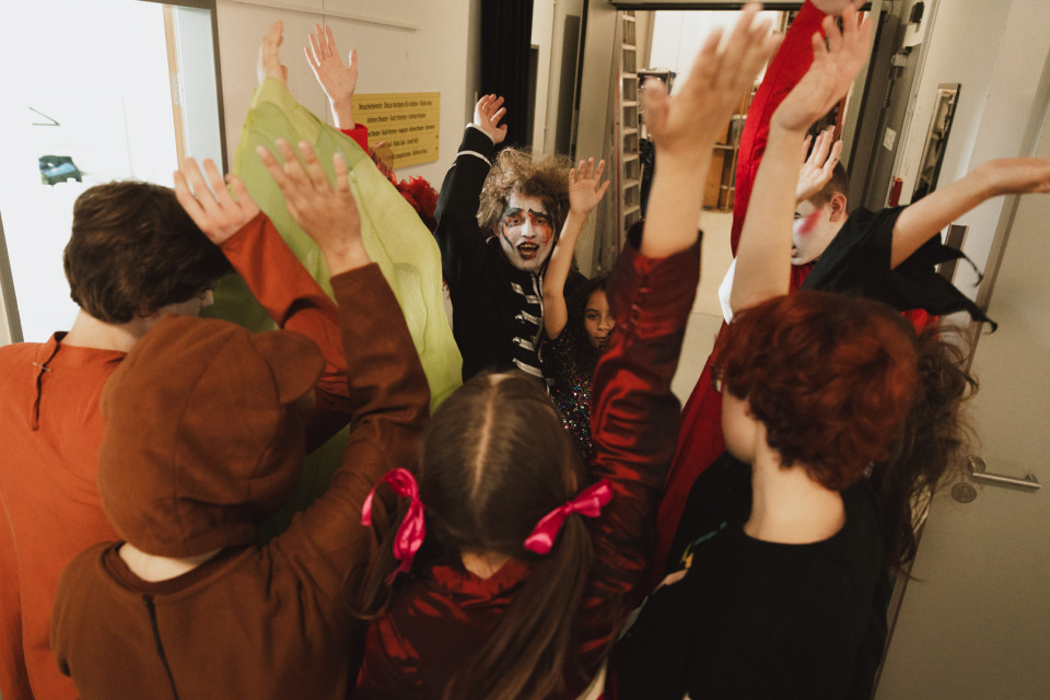 Eine Gruppe von verkleideten Kindern steht in einem Raum dicht beieinander und streckt die Arme nach oben. Einige tragen geschminkte Gesichter und bunte Kostüme, als würden sie feiern oder spielen.