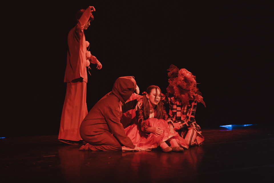 Auf einer dunkel beleuchteten Bühne sitzen und knien mehrere kostümierte Darsteller in rotem Licht um eine junge Frau in einem rosa Kleid. Die Szene wirkt dramatisch und theatralisch, mit fantasievollen Tier- oder Blumenmasken und einer weiteren Figur, die links stehend eine Geste ausführt.