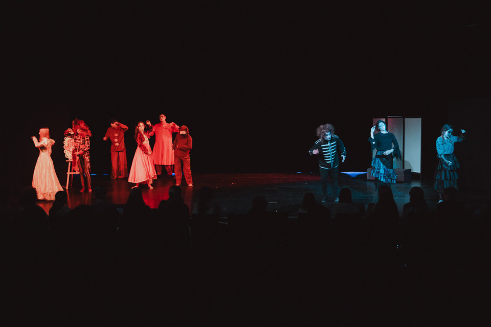 Auf einer dunklen Theaterbühne stehen mehrere kostümierte Darstellerinnen und Darsteller in rotem und blauem Licht verteilt. Im Vordergrund sind die Silhouetten des Publikums zu sehen, während die Szene wie eine dramatische Aufführung oder Tanzsequenz wirkt.