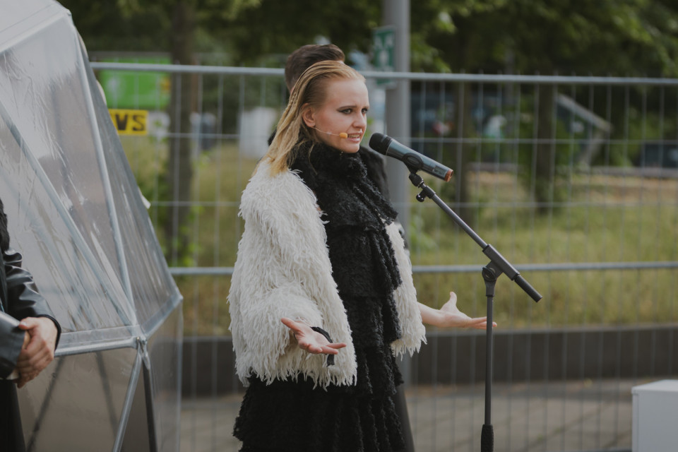 Eine Frau in einem schwarz-weißen, flauschigen Outfit steht draußen vor einem Mikrofon und scheint zu sprechen oder zu singen. Im Hintergrund sind ein Metallzaun, Grünflächen und weitere Personen teilweise zu sehen.