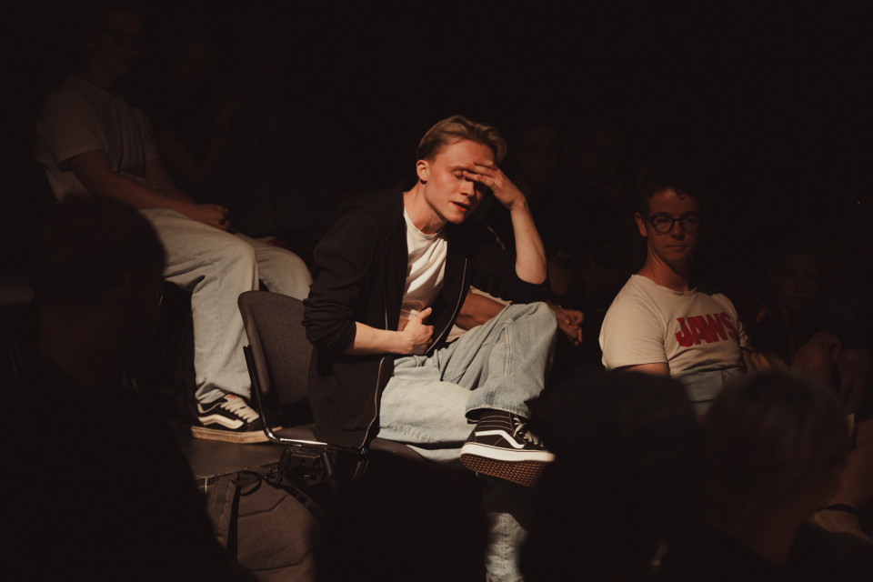 Ein junger Mann sitzt im dunklen Publikum im Scheinwerferlicht auf einem Stuhl und hält sich die Hand an die Stirn. Um ihn herum sitzen weitere Personen im Halbdunkel, wodurch eine konzentrierte, theaterartige Atmosphäre entsteht.