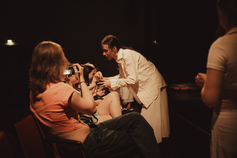 In einem dunklen Theaterraum beugt sich eine Person in weißer Kleidung zu sitzenden Zuschauerinnen und reicht ihnen offenbar etwas. Eine Frau im Vordergrund fotografiert oder filmt die Szene mit ihrem Smartphone, während rechts eine weitere Person unscharf im Bild steht.
