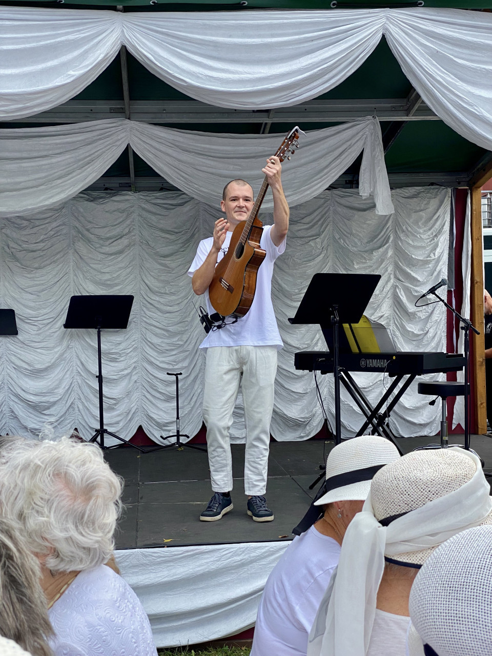 Ein Musiker steht auf einer Bühne vor weißen Stoffbahnen und hält eine Gitarre aufrecht in die Höhe. Im Vordergrund sitzen Zuschauer mit hellen Hüten, während neben dem Musiker Notenständer, Mikrofone und ein Yamaha-Keyboard zu sehen sind.