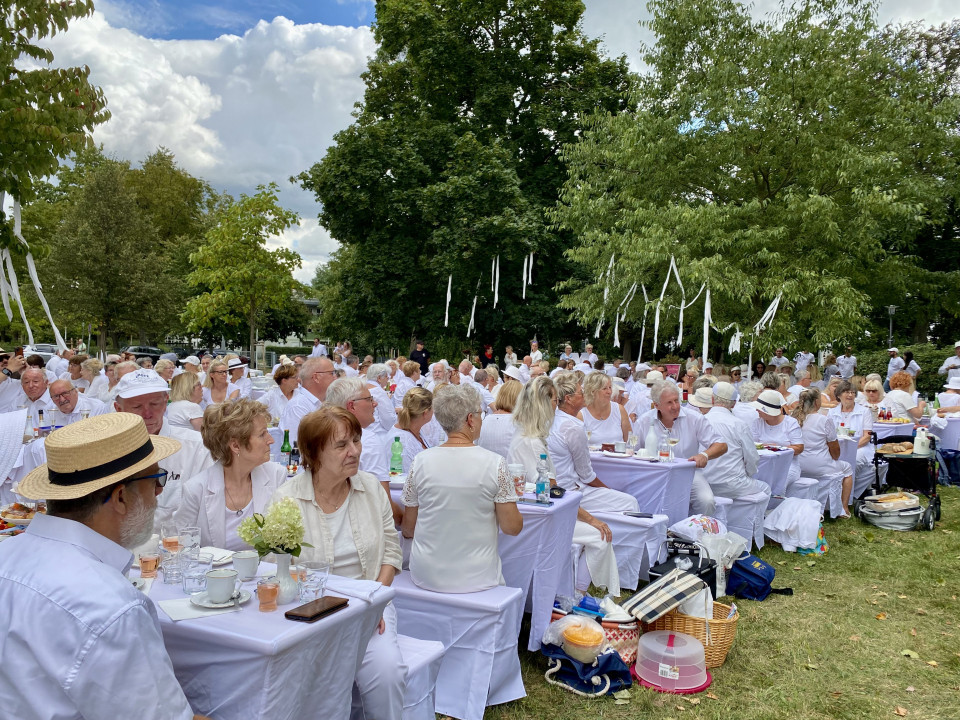 Viele Menschen sitzen bei einer festlichen Veranstaltung im Park an weiß gedeckten Tischen und tragen überwiegend weiße Kleidung. Zwischen den Bäumen hängen weiße Bänder, während unter einem bewölkten Himmel gemeinsam gegessen und getrunken wird.