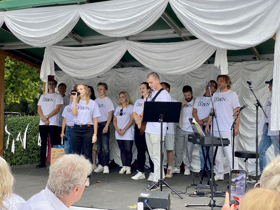 Eine Gruppe von Sängerinnen und Sängern in weißen T-Shirts steht auf einer geschmückten Bühne mit weißen Stoffbahnen. Eine blonde Frau singt ins Mikrofon, während ein Mann mit Gitarre und weitere Musiker neben Keyboard und Mikrofonständern bereitstehen.