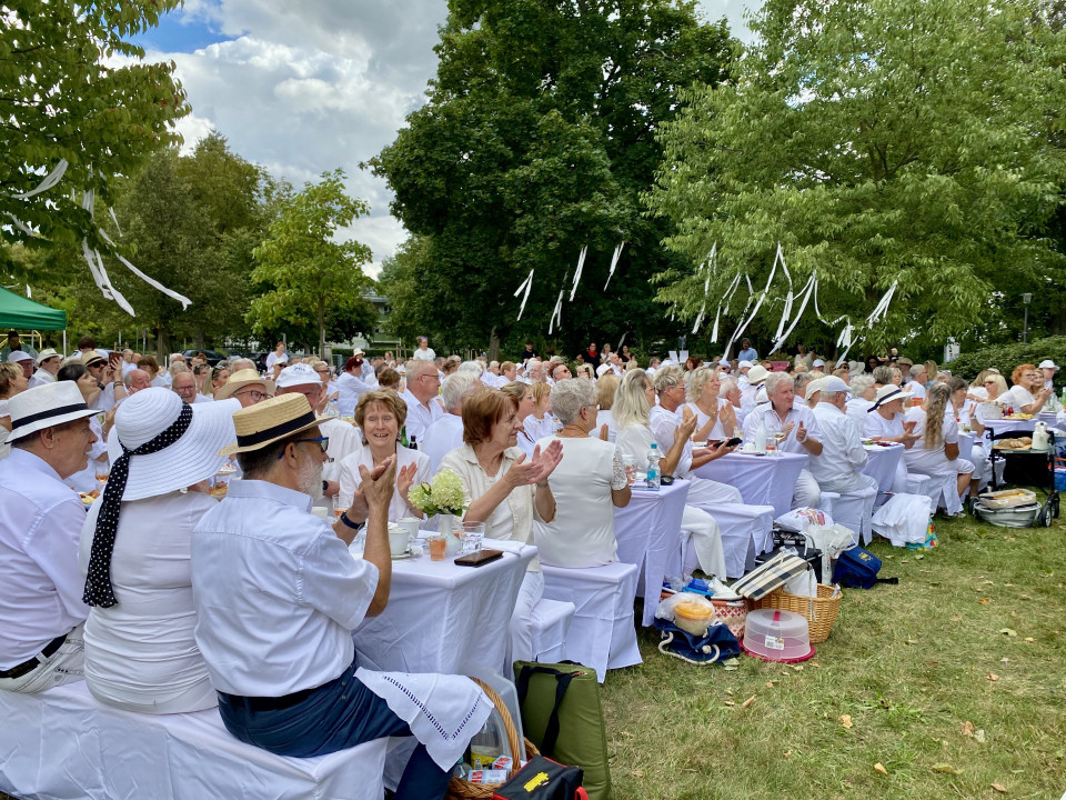 Viele Menschen in weißer Kleidung sitzen an langen, weiß gedeckten Tischen bei einem Fest im Park. Zwischen den grünen Bäumen hängen weiße Bänder, während die Gäste applaudieren und gemeinsam essen.
