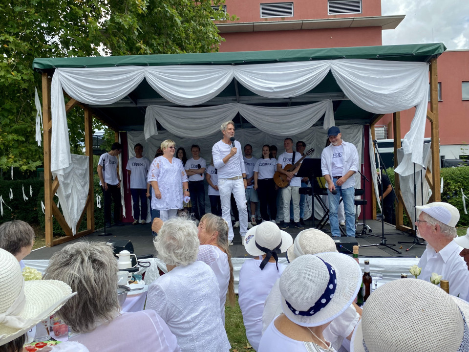 Auf einer dekorierten Freiluftbühne stehen mehrere weiß gekleidete Personen, während ein Mann mit Mikrofon zum Publikum spricht. Vor der Bühne sitzen zahlreiche Zuschauerinnen und Zuschauer in heller Kleidung und weißen Hüten an Tischen.