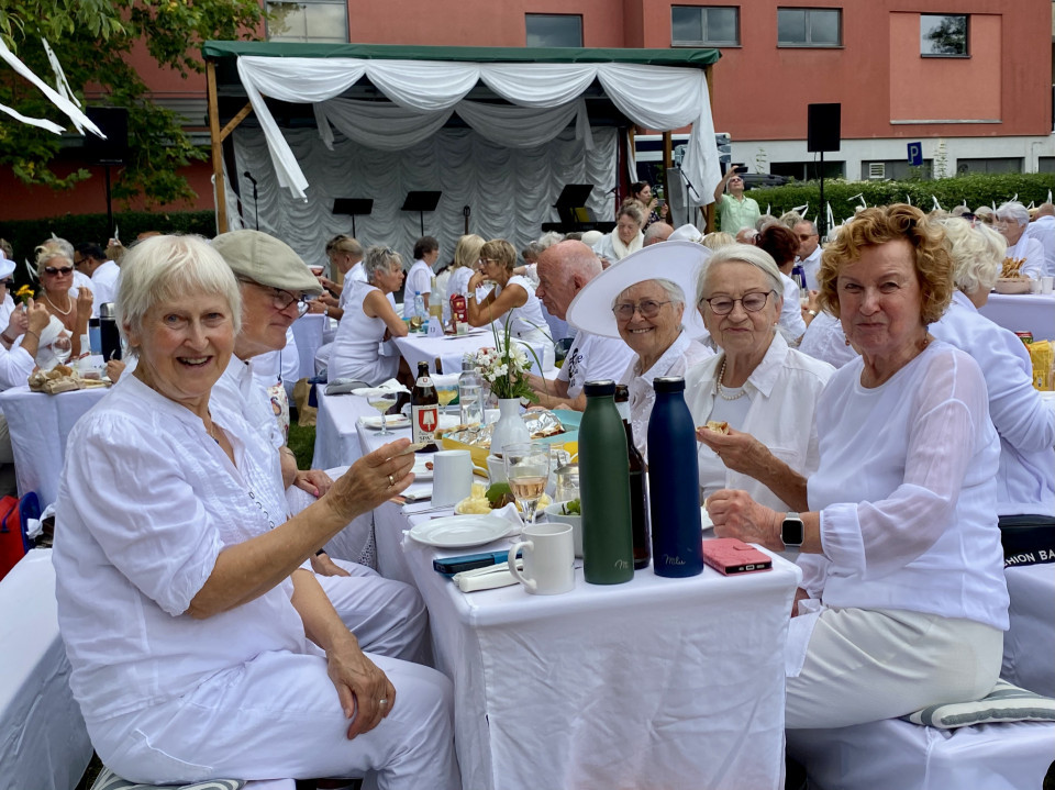 Eine Gruppe älterer Menschen in weißer Kleidung sitzt bei einer festlich gedeckten Tafel im Freien und lächelt in die Kamera. Im Hintergrund sind weitere Gäste, eine kleine Bühne mit weißen Vorhängen und ein rotes Gebäude zu sehen.