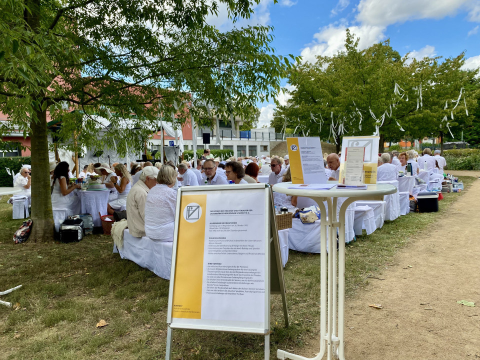 In einem Park sitzen viele Menschen ganz in Weiß gekleidet an langen Tischen unter Bäumen, während weiße Bänder als Dekoration in den Zweigen hängen. Im Vordergrund stehen Informationstafeln und ein Stehtisch, daneben führt ein Sandweg an der festlichen Veranstaltung vorbei.