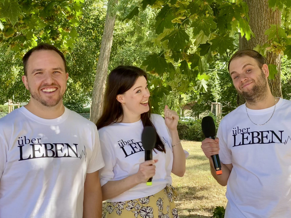 Drei lächelnde Personen stehen in einem grünen Park unter Bäumen und tragen weiße T-Shirts mit der Aufschrift „über LEBEN“. Zwei von ihnen halten Mikrofone in der Hand, während die Frau in der Mitte lachend zu dem Mann rechts schaut.