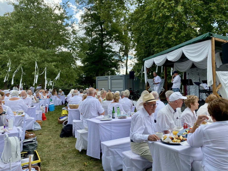 Viele Menschen in weißer Kleidung sitzen im Freien an weiß gedeckten Tischen in einem grünen Park. Rechts befindet sich eine dekorierte Bühne, auf der zwei Männer vor dem Publikum sprechen.