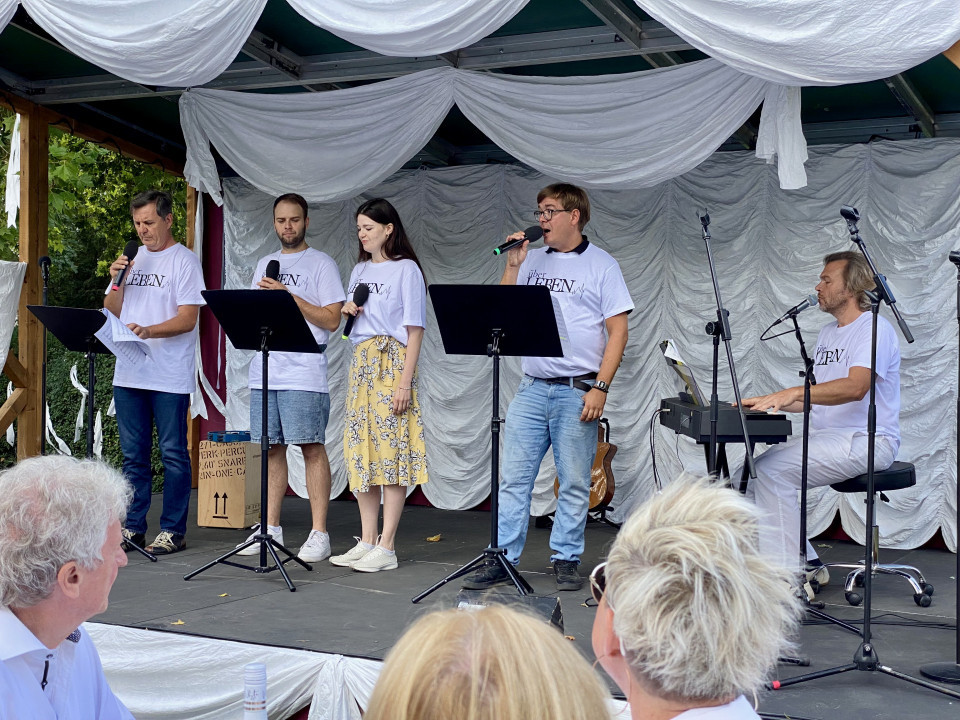 Eine Gruppe von Sängerinnen und Sängern steht auf einer dekorierten Bühne und tritt mit Mikrofonen vor Publikum auf. Rechts begleitet ein Musiker die Darbietung am Keyboard, während weiße Stoffbahnen den Bühnenhintergrund schmücken.