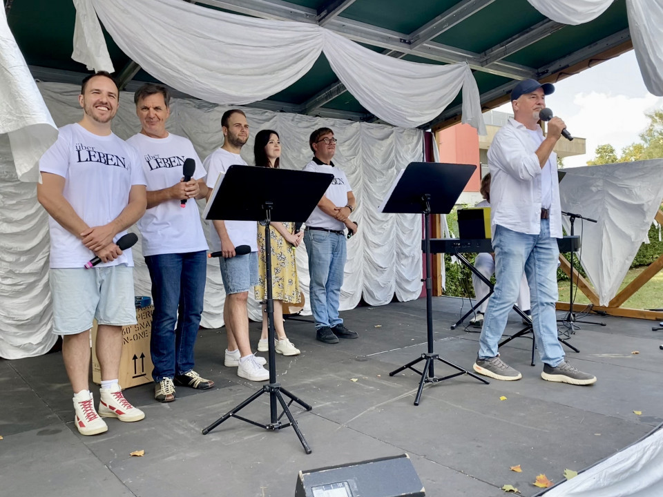 Auf einer Bühne stehen mehrere Menschen in weißen T-Shirts mit Mikrofonen, während ein Mann rechts in ein Mikrofon spricht. Die Bühne ist mit weißen Stoffbahnen dekoriert, und vor der Gruppe stehen Notenständer sowie ein Keyboard im Hintergrund.