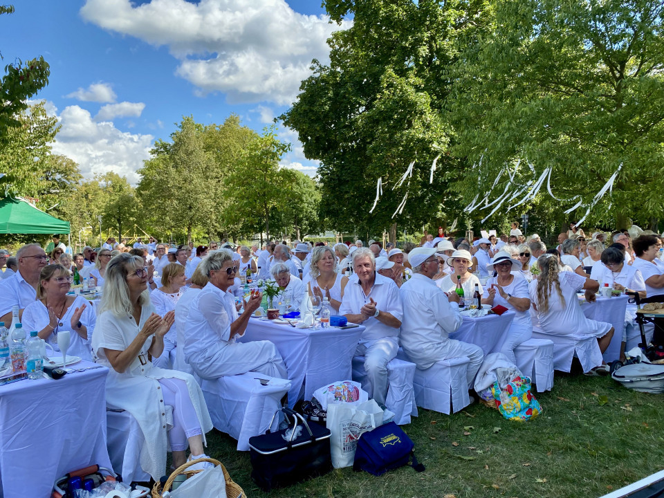 Eine große Gruppe überwiegend weiß gekleideter Menschen sitzt an langen, gedeckten Tischen in einem grünen Park. Über ihnen hängen weiße Bänder in den Bäumen, während ein blauer Himmel mit Wolken für eine sommerliche Atmosphäre sorgt.