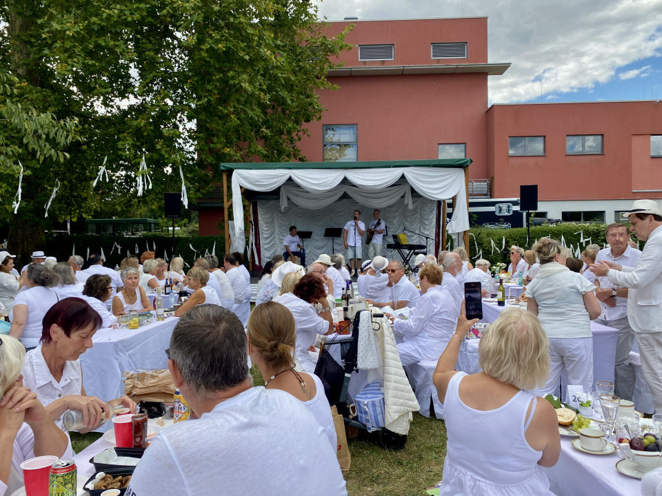 Viele Menschen in weißer Kleidung sitzen im Freien an langen gedeckten Tischen und schauen auf eine kleine Bühne mit Musikern. Im Hintergrund stehen ein rotes Gebäude, große Bäume mit weißen Bändern und ein bewölkter Himmel.