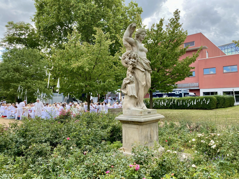 Eine steinerne Frauenstatue mit Blumen steht in einem üppigen Garten zwischen Rosen und grünen Bäumen. Im Hintergrund sitzen viele weiß gekleidete Menschen bei einer Veranstaltung vor einem roten Gebäude unter bewölktem Himmel.