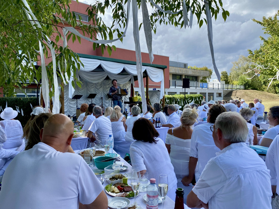 Viele weiß gekleidete Menschen sitzen draußen an gedeckten Tischen und schauen einer Musikaufführung auf einer weiß dekorierten Bühne zu. Über den Köpfen hängen lange weiße Bänder in den Bäumen, während im Hintergrund Gebäude und ein bewölkter Himmel zu sehen sind.