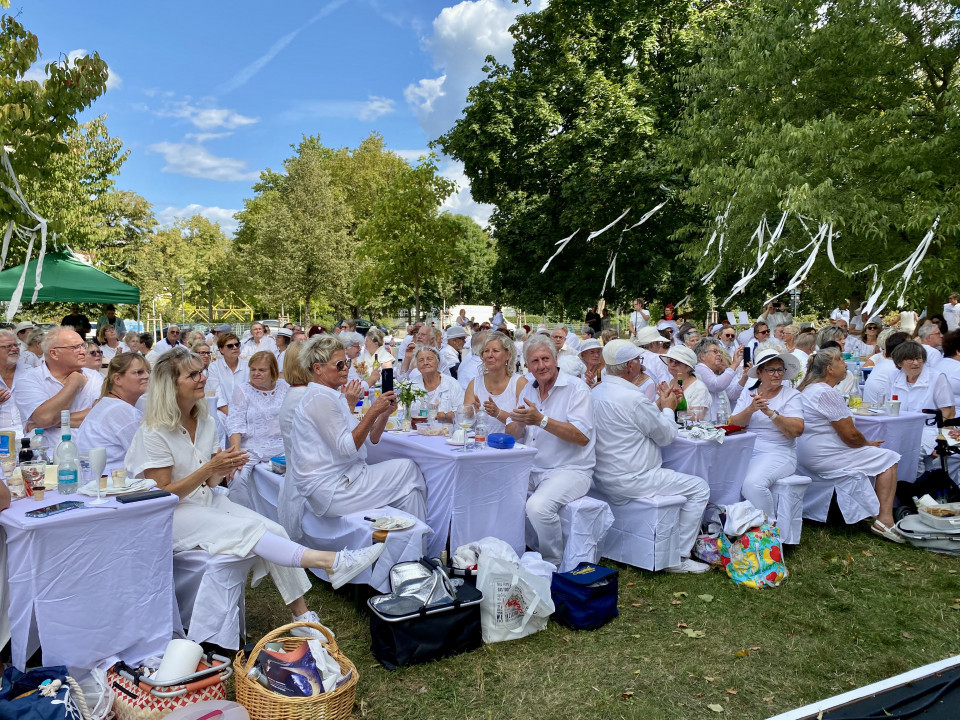 Viele Menschen in weißer Kleidung sitzen an langen, weiß gedeckten Tischen bei einem Fest im Park. Zwischen großen grünen Bäumen hängen weiße Bänder, während die Gäste essen, trinken und applaudieren.