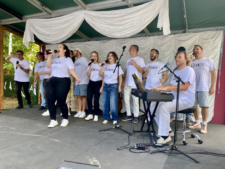 Eine Gruppe von Sängerinnen und Sängern in weißen T-Shirts steht auf einer Bühne und singt fröhlich in Mikrofone. Rechts begleitet ein Musiker die Gruppe am Keyboard, während weiße Stoffbahnen die Bühne dekorieren.