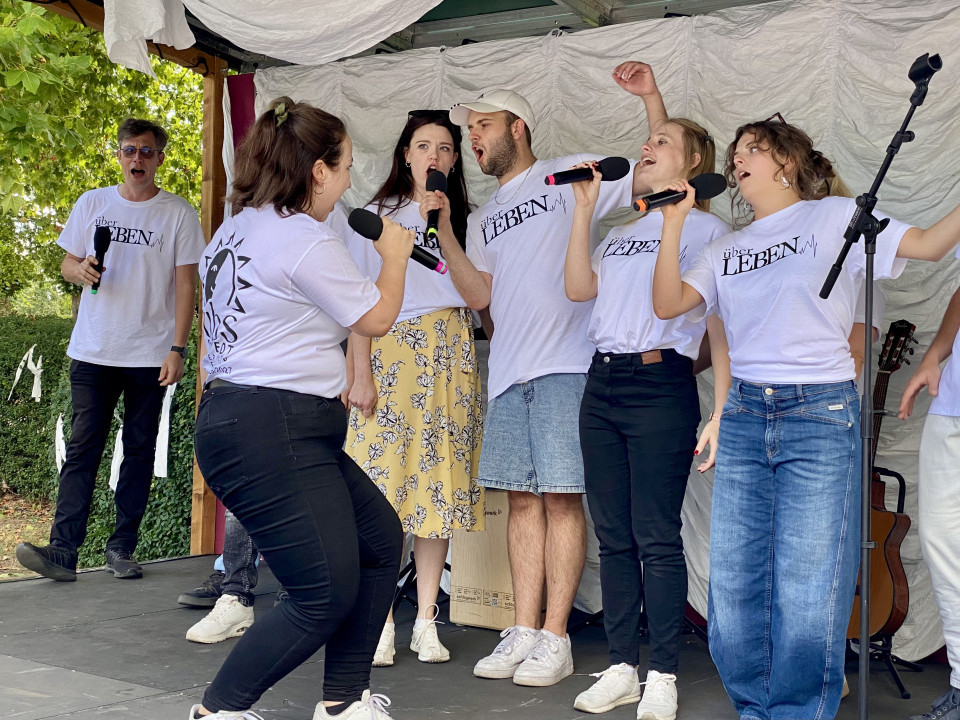 Eine Gruppe von Sängerinnen und Sängern steht auf einer kleinen Bühne im Freien und singt mit Mikrofonen. Alle tragen weiße T-Shirts mit der Aufschrift „über LEBEN“, im Hintergrund sind weiße Stoffbahnen, Bäume und eine Gitarre zu sehen.
