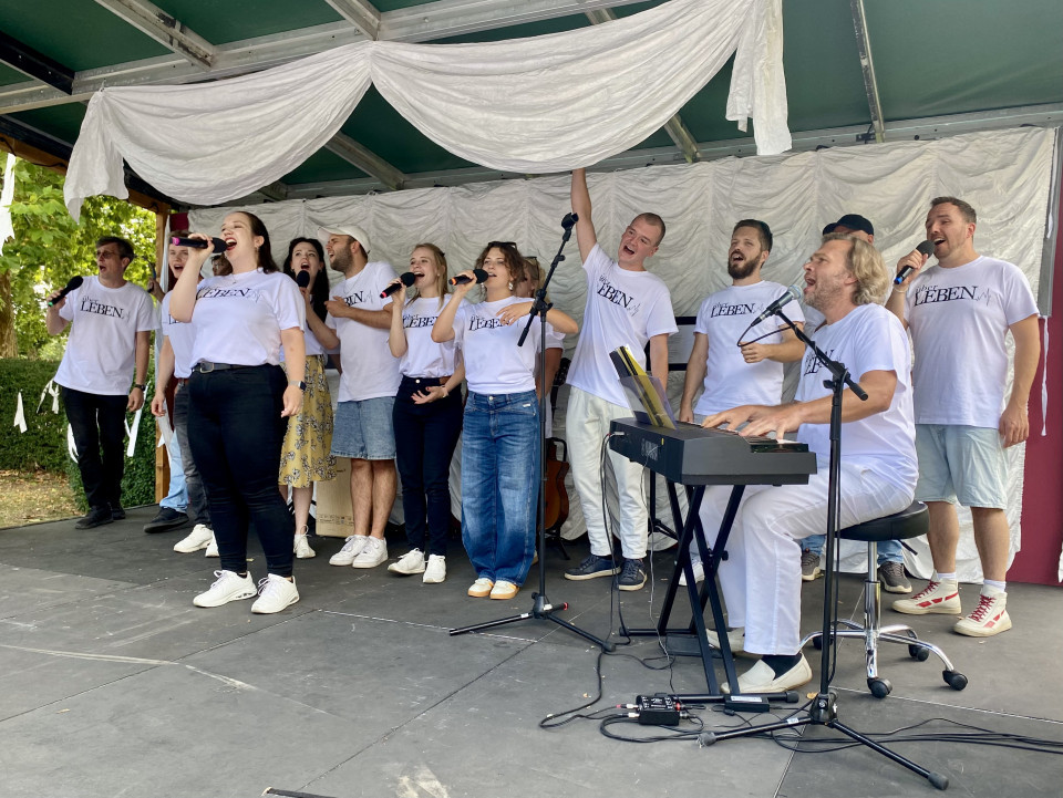 Eine Gruppe von Sängerinnen und Sängern in weißen T-Shirts tritt auf einer kleinen Bühne auf und singt mit Mikrofonen. Rechts begleitet ein Mann am Keyboard die Gruppe, während weiße Stoffbahnen die Bühne schmücken.