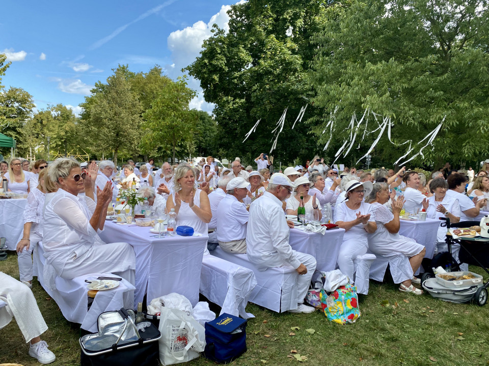 Eine große Gruppe überwiegend weiß gekleideter Menschen sitzt an festlich gedeckten Tischen im Grünen und applaudiert. Über ihnen hängen weiße Bänder in den Bäumen, während der Himmel blau und sonnig ist.