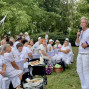 Ein Mann in weißer Kleidung spricht mit einem Mikrofon zu einer Gruppe von Menschen, die an weiß gedeckten Tischen im Freien sitzen. Die Veranstaltung findet in einem grünen Park statt, und viele Gäste tragen ebenfalls weiße Kleidung.