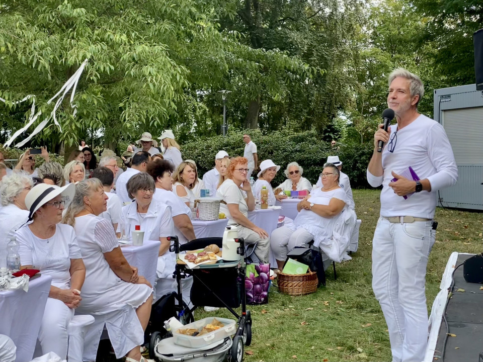 Ein Mann in weißer Kleidung spricht mit einem Mikrofon zu einer Gruppe von Menschen, die an weiß gedeckten Tischen im Freien sitzen. Die Veranstaltung findet in einem grünen Park statt, und viele Gäste tragen ebenfalls weiße Kleidung.