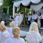 Ein Mann spricht mit einem Mikrofon vor einem Publikum, während er auf einer weiß dekorierten Freiluftbühne ein kleines Heft hochhält. Neben ihm stehen mehrere Personen in weißen T-Shirts, und die Gäste im Vordergrund sitzen ebenfalls überwiegend in Weiß gekleidet an Tischen.