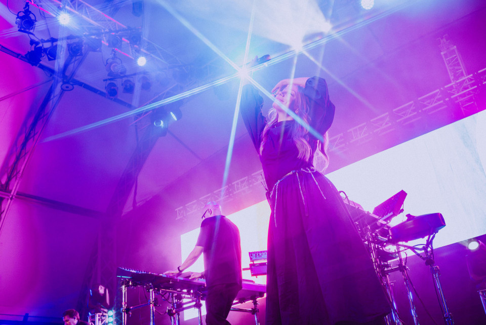 Eine Sängerin steht auf einer Bühne und hebt unter grellen blau-violetten Scheinwerfern die Arme. Im Hintergrund spielen Musiker an elektronischen Instrumenten vor einer großen hellen Leinwand.