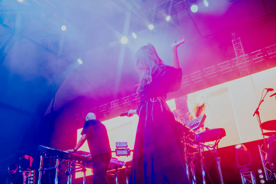 Eine Sängerin steht mit erhobener Hand auf einer Konzertbühne, während Musiker an elektronischen Instrumenten spielen. Bunte pinke und blaue Lichter sowie eine große LED-Wand erzeugen eine atmosphärische Showkulisse.