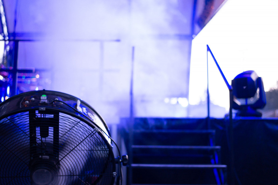 Im Vordergrund steht ein großer Ventilator, der von blau-violettem Licht angestrahlt wird. Im Hintergrund sind unscharf eine Treppe, Geländer und ein Bühnenscheinwerfer in einer atmosphärischen Veranstaltungsszene zu sehen.