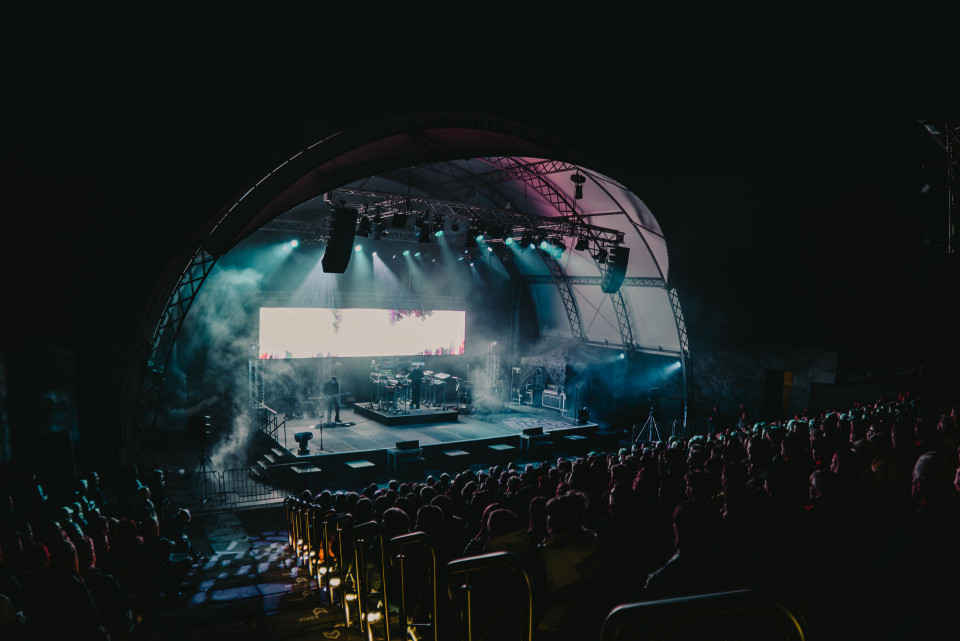 Auf einer großen Freilichtbühne findet nachts ein Konzert mit hellen Scheinwerfern, Nebel und einer leuchtenden Bildschirmwand statt. Vor der Bühne sitzt und steht ein großes Publikum im dunklen Zuschauerbereich.